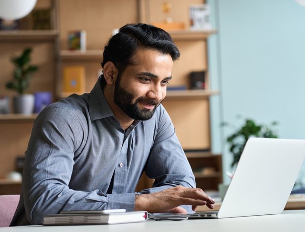 Smiling indian business man working on laptop at home office. Young indian student or remote teacher using computer remote studying, virtual training, watching online education webinar at home office.