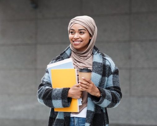 Female student in hijab and coat with books and cup of coffee ready to study.