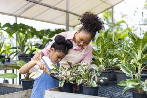 a black woman in a green house helps a small black child water a plant